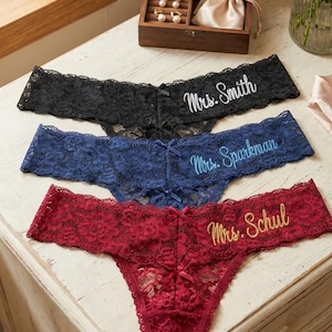 May include: Three pairs of personalised lace underwear in black, navy blue, and burgundy. Each pair is embroidered with "Mrs." and a surname in a contrasting colour. A wooden jewellery box and a vase of roses are in the background.