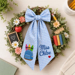 Könnte beinhalten: Ein Weinrebenkranz dekoriert mit grünem Blattwerk, weißen und rosa Blumen, einem roten Apfel, einer kleinen Tafel mit Herz, einem Holzlineal, Miniaturbüchern und einer großen blau-weißen Vichy-Schleife mit Rucksack und dem Text Miss Chloe.