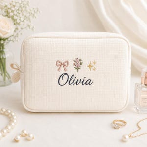 Könnte beinhalten: Ein rechteckiges Etui aus cremefarbenem, strukturiertem Stoff mit einer Stickerei aus einer rosa Schleife, einem kleinen Blumenstrauß und goldenen Sternen sowie dem Namen Olivia in dunkelblauer Schreibschrift auf der Vorderseite.