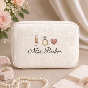 May include: A rectangular off-white textured fabric pouch featuring embroidered icons of a champagne flute, a diamond ring, and a heart. Below the icons, the text Mrs. Parker is embroidered in a dark grey cursive script font.