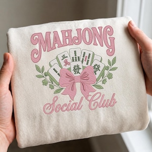 May include: Cream-colored sweatshirt with a pink embroidered design. The design features the word "MAHJONG" above a collection of Mahjong tiles, a pink bow, and the words "Social Club". Green leaves and floral accents surround the tiles.