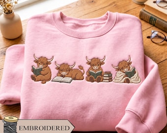 May include: A light pink crewneck sweatshirt featuring an embroidered design of four brown Highland cattle characters. Each character is depicted in a different pose, with three reading books and one lying down with an open book. The text EMBROIDERED is visible.