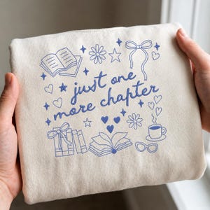 May include: A cream-colored tote bag with blue illustrations of books, flowers, hearts, stars, and a coffee cup. The text "just one more chapter" is written in blue script. The bag is held by two hands.