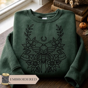 May include: A forest green crewneck sweatshirt featuring a black embroidered design of a moth with a skull-shaped body, surrounded by floral motifs and a crescent moon symbol centered above the moth's head, with long sleeves and ribbed cuffs.