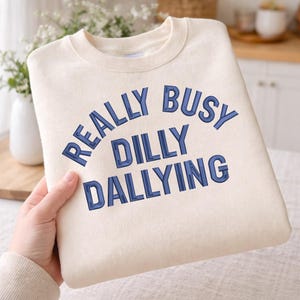 May include: Cream-colored sweatshirt with the embroidered text "REALLY BUSY DILLY DALLYING" in blue. The sweatshirt is folded and held by a person. The background includes a vase of flowers and a kitchen setting.