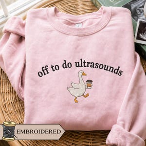 May include: A light pink crewneck sweatshirt featuring black embroidered text that reads off to do ultrasounds, accompanied by an embroidered graphic of a white goose with an orange beak and feet, holding a brown coffee cup in its wing.