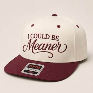 May include: A cream-colored snapback cap featuring the embroidered text I COULD BE Meaner in dark maroon. The cap has a flat maroon brim and a sticker on the brim that reads OTTO PREMIUM FABRIC S-N-A-P ONE SIZE FITS MOST SNAPBACK CLOSURE.
