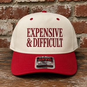 May include: A five-panel baseball cap with an off-white crown and a red brim. The front features red embroidered text that reads EXPENSIVE & DIFFICULT. A silver and black sticker on the brim displays the text OTTO PREMIUM FABRIC 5 PANEL MID PROFILE.