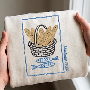 Può includere: Felpa color crema con un disegno ricamato. Mostra un cestino di baguette, due pesci e il testo "Matthew 14:20" in blu. Il disegno è incorniciato da un bordo blu. La felpa è tenuta da mani.