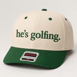 May include: A cream-colored baseball cap with a dark green brim and matching green embroidered text on the front that reads he's golfing. The cap features a small green button on top and a rectangular sticker on the brim with the text OTTO.