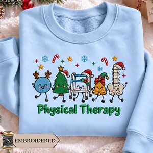 May include: A light blue crewneck sweatshirt featuring an embroidered graphic of five festive characters including a blue sphere with antlers, a Christmas tree, a walker, a bell, and a spine, all wearing Santa hats, with the text Physical Therapy.