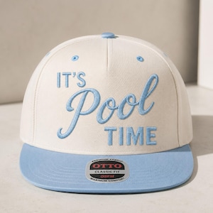 Op de afbeelding: Een crèmekleurige baseballpet met een platte lichtblauwe klep. De voorkant is voorzien van geborduurd lichtblauw tekst die luidt IT'S Pool TIME. Een ronde sticker op de klep toont de tekst OTTO PREMIUM FABRIC CLASSIC FIT OSFM MAXIMUM COMFORT.