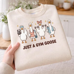 May include: Cream-colored sweatshirt featuring a cartoon design of four geese engaged in various gym activities. The geese are depicted with weights, a yoga mat, and water bottles. The text "JUST A GYM GOOSE" is printed below the geese.