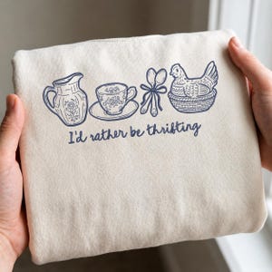 Könnte beinhalten: Cremefarbene Tragetasche mit einer blauen Strichzeichnung einer Kanne, einer Teetasse, Löffeln und einem Huhn in einem Korb. Der Text "I'd rather be thrifting" ist darunter gedruckt.