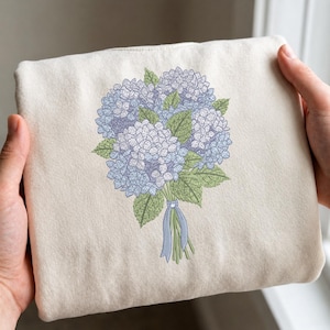 May include: A cream-colored tote bag featuring a detailed embroidered bouquet of blue and purple hydrangeas with green leaves. The flowers are tied with a blue ribbon. The bag is held by two hands.