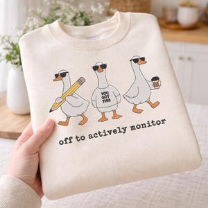 May include: Cream-colored sweatshirt featuring three cartoon geese wearing sunglasses. One goose holds a pencil, another has "YOU GOT THIS" on its shirt, and the third carries a coffee cup. The text "off to actively monitor" is printed below.