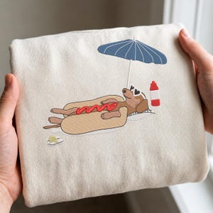 May include: A cream-colored tote bag featuring an embroidered design of a dachshund relaxing on a hot dog, under a blue umbrella. The dog wears heart-shaped sunglasses, with a ketchup bottle nearby. A fun, summer-themed design.
