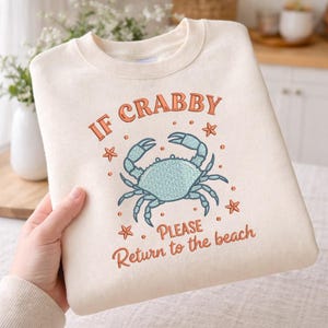 May include: Cream-colored sweatshirt with the text "IF CRABBY PLEASE Return to the beach" in orange script. A blue crab illustration is centered below the text, with orange starfish accents. The sweatshirt is folded.