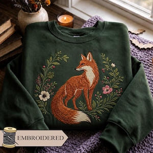 May include: A dark forest green crewneck sweatshirt featuring a detailed embroidered design of a red fox surrounded by a wreath of green leaves, small red berries, and white and pink flowers. The word EMBROIDERED is printed on a small tag.