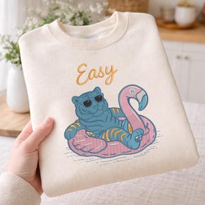 Puede incluir: Sudadera color crema con un tigre azul con gafas de sol, relajándose en un flotador de flamenco rosa. La palabra "Easy" está escrita encima del tigre en letras doradas. Diseño divertido y casual.