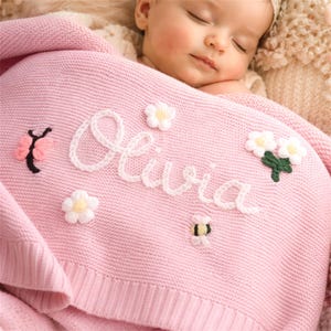 Pode incluir: Um cobertor de bebê rosa de malha com o nome "Olivia" bordado em branco. O cobertor é decorado com flores brancas, uma borboleta e uma abelha. O bebê está dormindo pacificamente.