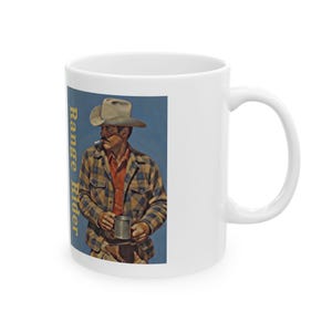 Op de afbeelding: Witte keramische mok met een vintage illustratie van een cowboy met een hoed, die een mok vasthoudt en een pijp rookt. De mok heeft de woorden "Range Rider" in gele tekst op een blauwe achtergrond.