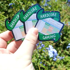 Può includere: Una collezione di adesivi con un design a foglio di calcolo e le parole "HARDCORE GAMING" su banner verdi. Gli adesivi hanno una finitura olografica e sono tenuti davanti a uno sfondo di fogliame verde e fiori blu.