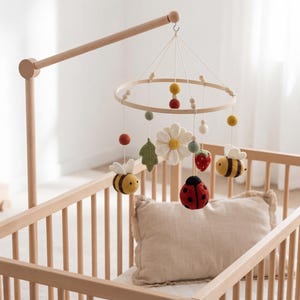 Puede incluir: Una cuna de madera con un móvil colgante. El móvil presenta abejas de fieltro, una mariquita, una margarita y otras formas coloridas. La cuna es de color claro y tiene una almohada suave.