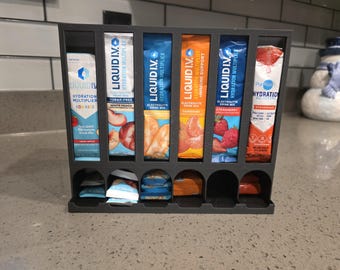 May include: A dark gray plastic organizer rack featuring six vertical slots holding various Liquid I.V. hydration multiplier drink mix packets, with additional individual packets stored in the rounded compartments at the base.