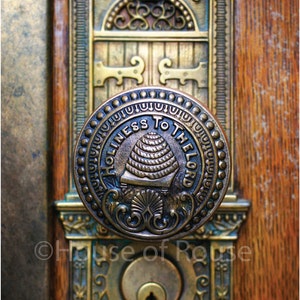 May include: Close-up of an antique door with a brass doorknob. The doorknob is round with the words "HOLINESS TO THE LORD" around a beehive design. The door has decorative brass accents and the years 1853 and 1893.