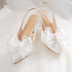 Puede incluir: Un par de zapatos de tacón blancos de satén con tiras traseras y puntera afilada. Cada zapato presenta un lazo grande y transparente con un detalle de perla. Los tacones se colocan sobre un fondo blanco texturizado, lo que sugiere un tema nupcial.