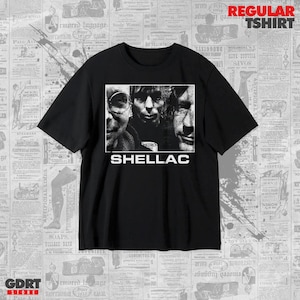 Puede incluir: Una camiseta negra de manga corta con un gráfico rectangular central que presenta una impresión fotográfica en blanco y negro de tres rostros. La palabra SHELLAC está impresa en letras mayúsculas blancas debajo de la imagen en el frente.