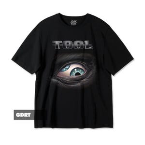 May include: Black t-shirt featuring the word "TOOL" in silver lettering above a detailed eye graphic with blue and green hues. The shirt has short sleeves and a crew neck.