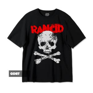 Puede incluir: Camiseta negra con un diseño de calavera y huesos cruzados blancos y la palabra "RANCID" en rojo. La camiseta tiene mangas cortas y cuello redondo. El logotipo de Good Print es visible en la esquina superior derecha.