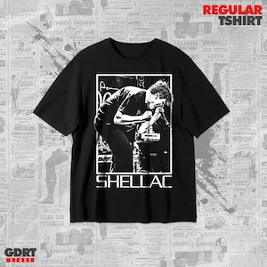 May include: A black short-sleeve t-shirt featuring a white graphic print of a person singing into a microphone on stage. The text SHELLAC is printed in white capital letters below the graphic. The shirt has a crew neckline and a standard fit.