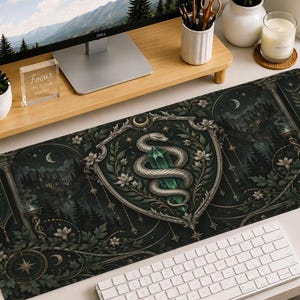 Könnte beinhalten: Eine rechteckige Schreibtischunterlage mit einer dunkelgrünen und schwarzen Illustration einer Schlange, die sich um einen grünen Kristall windet, zentriert in einem verzierten Schild. Das Design zeigt florale Motive, Monde und Sterne.