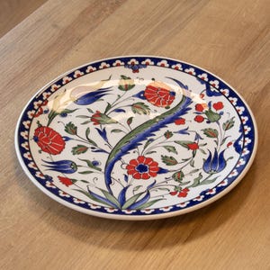 May include: White ceramic plate with a blue border and floral design. The plate features red flowers, blue tulip-like shapes, and green leaves. The design is centered on the plate and is set against a white background.