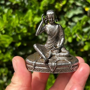 May include: A silver-colored figurine of a seated figure in a meditative pose, holding a small bowl. The figure is on a decorative base with ornate detailing. The figurine is held in a hand against a blurred green background.