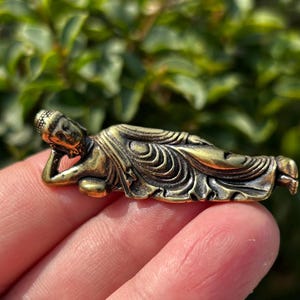 Op de afbeelding: Een kleine, bronzen Boeddha-beeld in een liggende houding. De figuur is gedetailleerd met ingewikkelde kledingplooien en gelaatstrekken. Het beeld wordt in een hand gehouden, met een vage groene achtergrond.