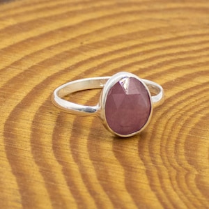 May include: A silver ring with a large, faceted, oval-shaped, pink gemstone. The ring is set on a wooden surface with visible wood grain. The gemstone has a slightly translucent appearance.