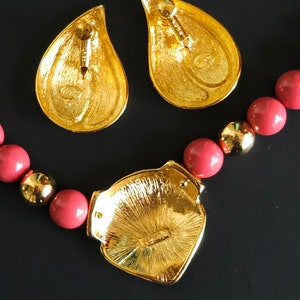 Pu&ograve; includere: Una collana dorata con un grande ciondolo a forma di cuore e una fila di perle color corallo. Il ciondolo presenta un design dettagliato e una piccola iscrizione sul retro. La collana &egrave; accompagnata da un paio di orecchini a goccia abbinati.