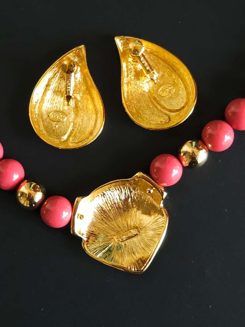 Pu&ograve; includere: Una collana dorata con un grande ciondolo a forma di cuore e una fila di perle color corallo. Il ciondolo presenta un design dettagliato e una piccola iscrizione sul retro. La collana &egrave; accompagnata da un paio di orecchini a goccia abbinati.