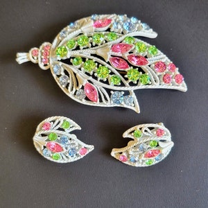 Pu&ograve; includere: Un set di gioielli in metallo bianco composto da una spilla e orecchini con un motivo a foglia. La spilla ha un ciondolo pendente e gli orecchini hanno la forma di foglie. I gioielli sono decorati con strass rosa, verdi e blu.