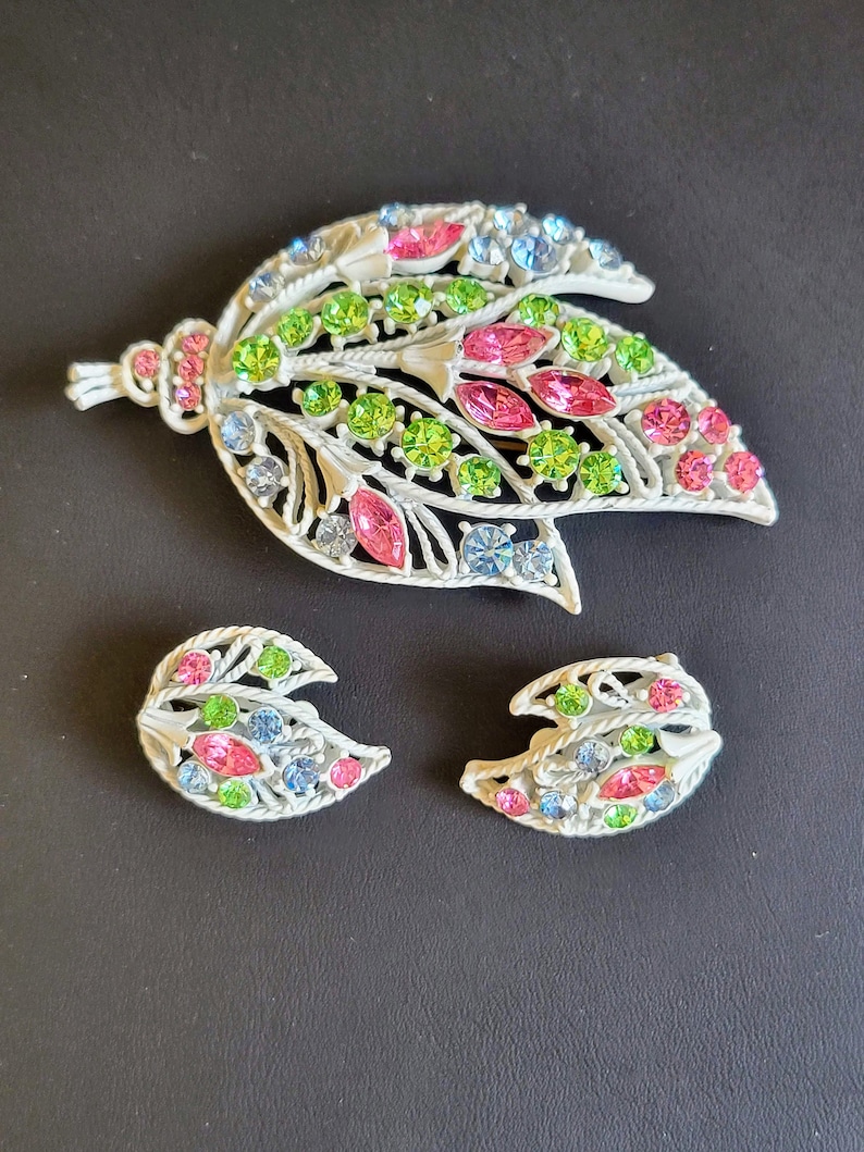Pu&ograve; includere: Un set di gioielli in metallo bianco composto da una spilla e orecchini con un motivo a foglia. La spilla ha un ciondolo pendente e gli orecchini hanno la forma di foglie. I gioielli sono decorati con strass rosa, verdi e blu.