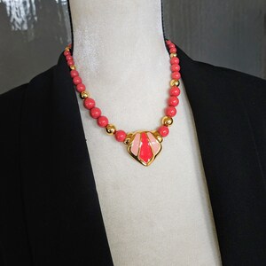 Pu&ograve; includere: Una collana d'oro con un ciondolo in smalto rosa e rosso. La collana &egrave; composta da perline rosse e distanziatori dorati.