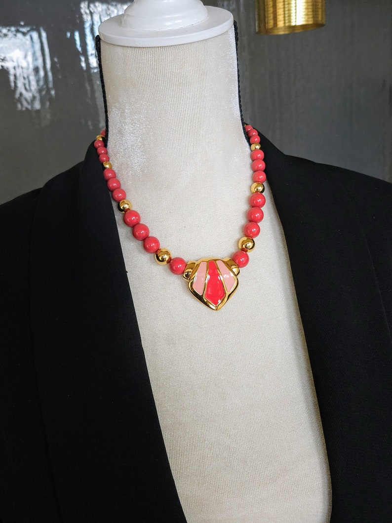 Pu&ograve; includere: Una collana d'oro con un ciondolo in smalto rosa e rosso. La collana &egrave; composta da perline rosse e distanziatori dorati.
