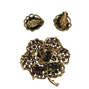 Pode incluir: Um broche dourado e brincos de press&atilde;o correspondentes com um design floral com strass. A parte de tr&aacute;s do broche e dos brincos s&atilde;o marcados com "Weiss".