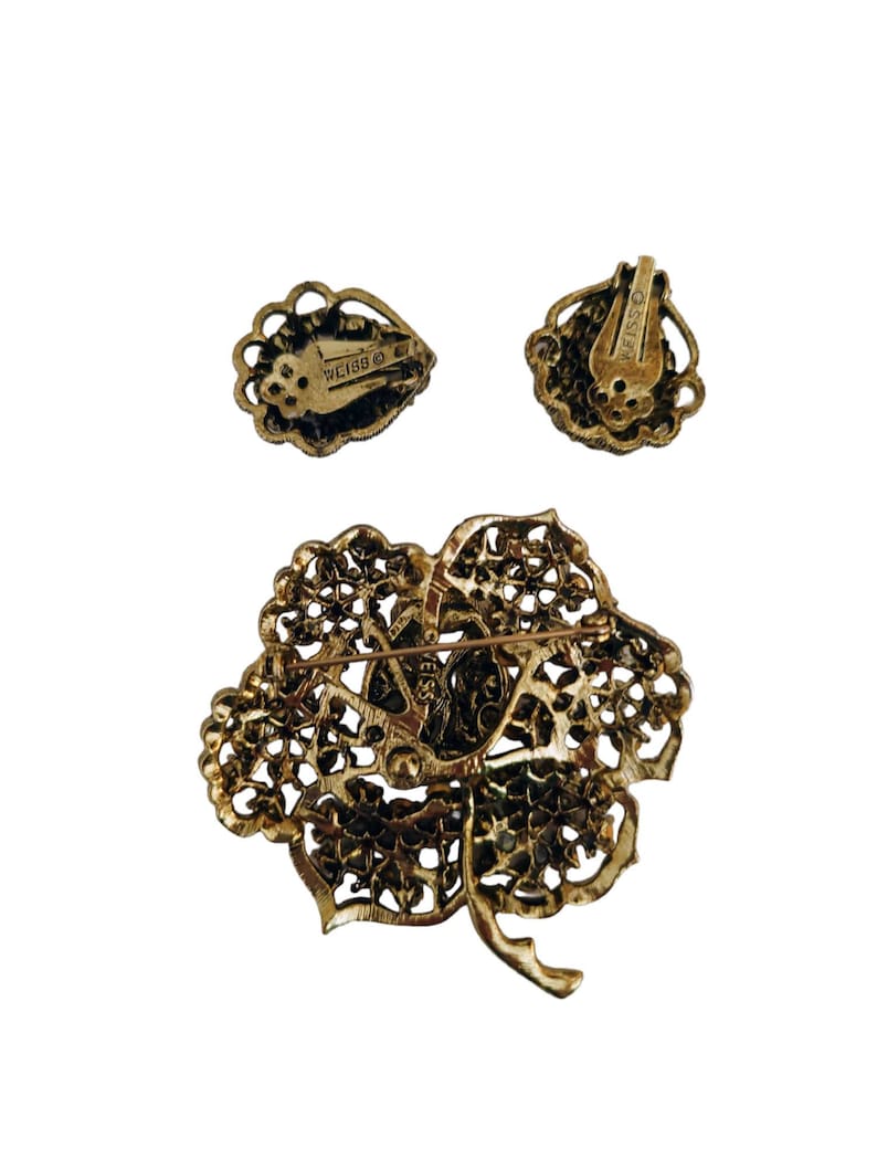 Pode incluir: Um broche dourado e brincos de press&atilde;o correspondentes com um design floral com strass. A parte de tr&aacute;s do broche e dos brincos s&atilde;o marcados com "Weiss".