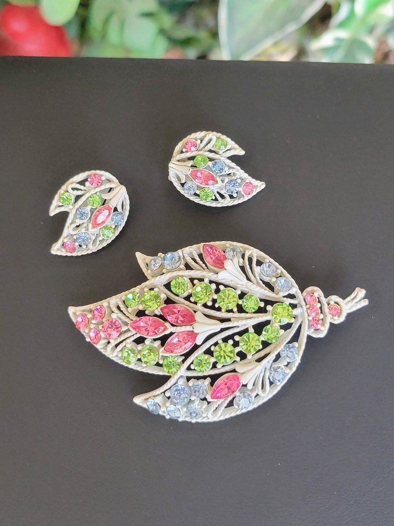 Pu&ograve; includere: Un set di tre spille a forma di foglia con strass in tonalit&agrave; rosa, verde e blu. Le spille sono realizzate in metallo argentato e hanno uno stile delicato e vintage.