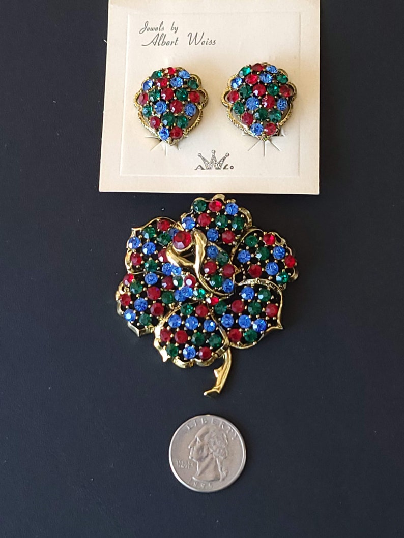 Pode incluir: Um conjunto de joias vintage de Albert Weiss. O conjunto inclui um broche e um par de brincos. O broche tem a forma de uma flor com pedras de imita&ccedil;&atilde;o vermelhas, azuis e verdes. Os brincos t&ecirc;m a forma de um cora&ccedil;&atilde;o e tamb&eacute;m apresentam pedras de imita&ccedil;&atilde;o vermelhas, azuis e verdes.